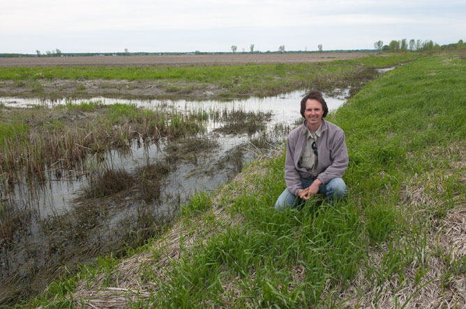 A man crouching in the grass at the edge of a stream
